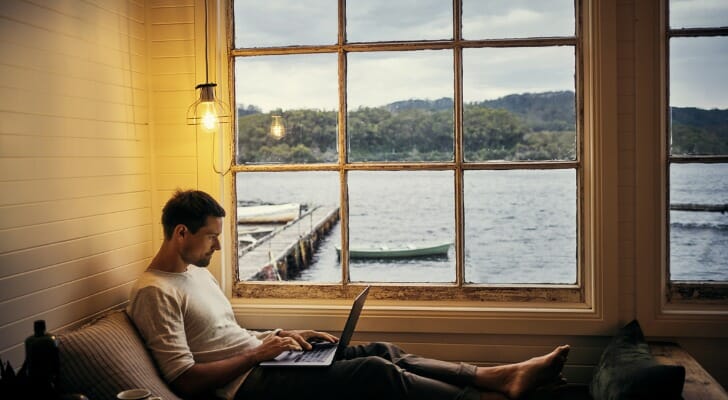 Image shows a person sitting at the window of their lakeside second home and working remotely on their laptop. SmartAsset crunched the numbers to conduct its latest study on the the hottest secondary home markets.
