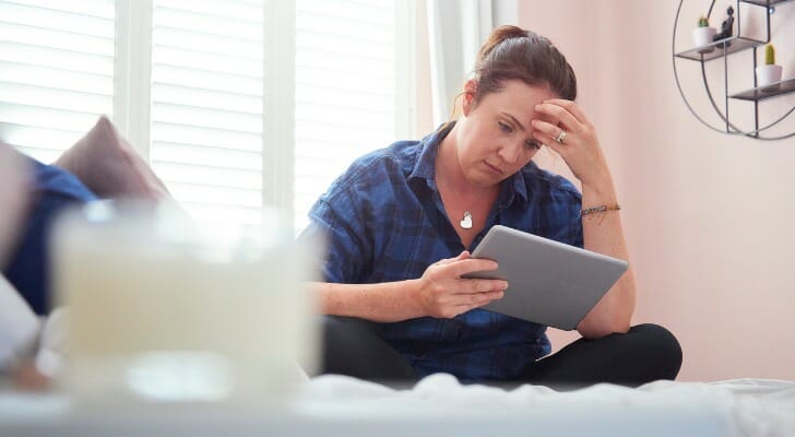 Image shows a person wearing a navy blue shirt and black pants sitting on a bed holding a tablet and rubbing their forehead as they look at their financial information. This study by SmartAsset finds the U.S. cities with the most financial stress.