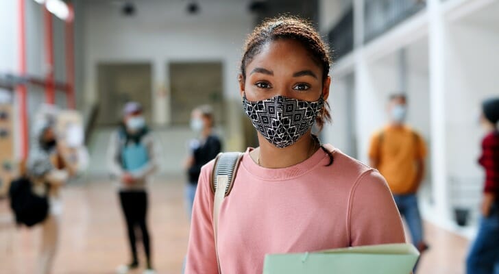 Image shows a student standing in a school hallway, holding notebooks and wearing a face mask to protect from COVID-19 and other viruses. SmartAsset analyzed various data sources to conduct its latest study on the top U.S. states for higher education. 