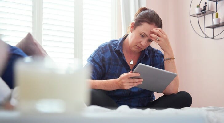 图像显示一个人穿着深蓝色衬衫和黑裤子坐在床上拿着平板电脑和擦额头上看他们的财务信息。本研究通过SmartAsse伟德ios appt发现最多的美国城市财政压力。