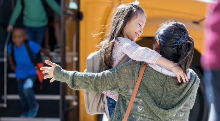 Image shows a young child wearing a light pink shirt and gray backpack hugging a guardian after school; another young child disembarks the yellow school bus in the background. SmartAsset analyzed data for 200 U.S. metro areas to find the best places to raise kids.