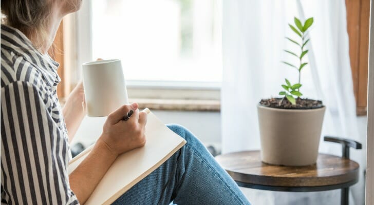 Image shows a person taking a break from work; they are sitting by a window and a potted plant, with a mug in one hand and a notebook and pen in the other. SmartAsset analyzed data to find the best cities for work-life balance.