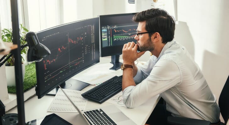 A man sits at a desk looking at two screens of financial market charts and graphs as he mulls what moves to make