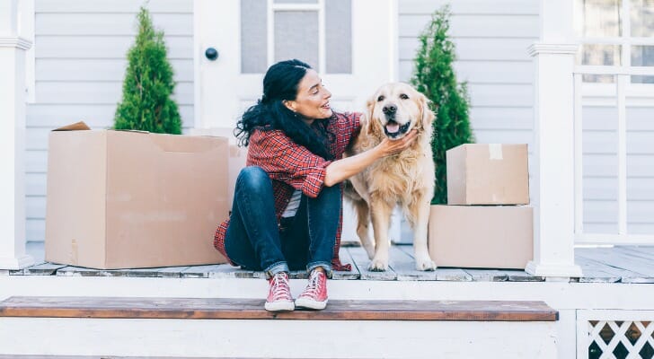 Images shows a young woman wearing a red plaid shirt, blue jeans and red sneakers sitting on white porch steps and petting an adult golden retriever. Moving boxes sit on either side of the woman and the dog. The dog is smiling. For this study, SmartAsset looked at data for 100 of the largest U.S. cities to find the most dog-friendly cities in the country.