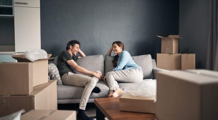 Image shows two new homeowners surrounded by unpacked boxes and sitting on a beige couch, talking happily. The couch sits on a wooden floor and against a gray wall. SmartAsset analyzed various data points in this study to find the best states for homeowners.