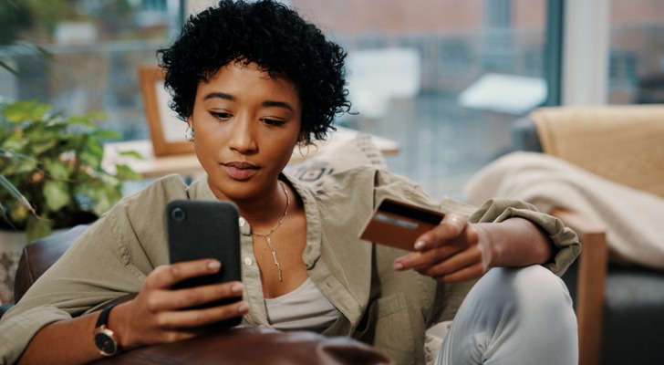 Image shows a woman holding a credit card in one hand while holding her phone in the other. SmartAsset analyzed various datasets to identify where credit card debt is becoming more manageable.