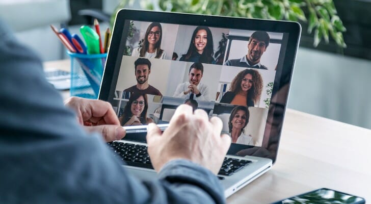 Image shows a person's hands at the keyboard of their laptop; their laptop screen shows a grid of faces in a video conference call. SmartAsset analyzed data to find the best cities to work from home in 2021.