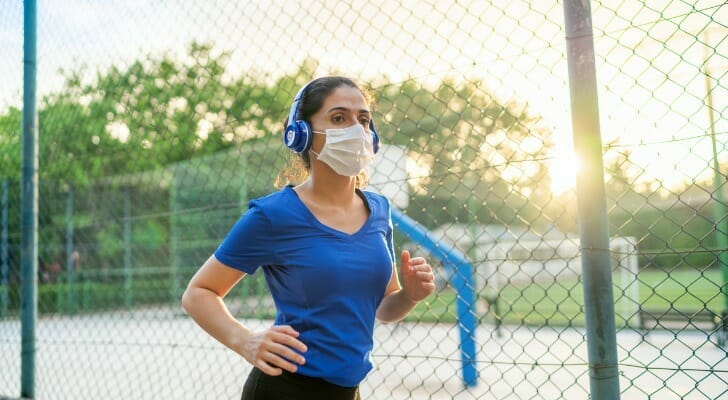 Image shows a young woman wearing a blue t-shirt, blue headphones, black leggings and a mask; she is jogging in a city park. SmartAsset analyzed data from various sources to find the best cities for runners.