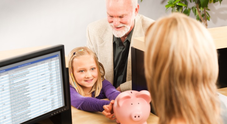 An older adult sits with a grandchild at the desk of a bank representative; the child holds a piggy bank. They are at the bank to ask about opening a trust bank account.